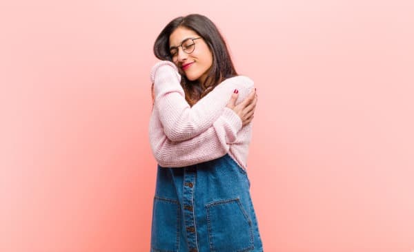 Woman with dark hair and glasses hugging herself with pink back drop, cosmetic dermatology in san antonio